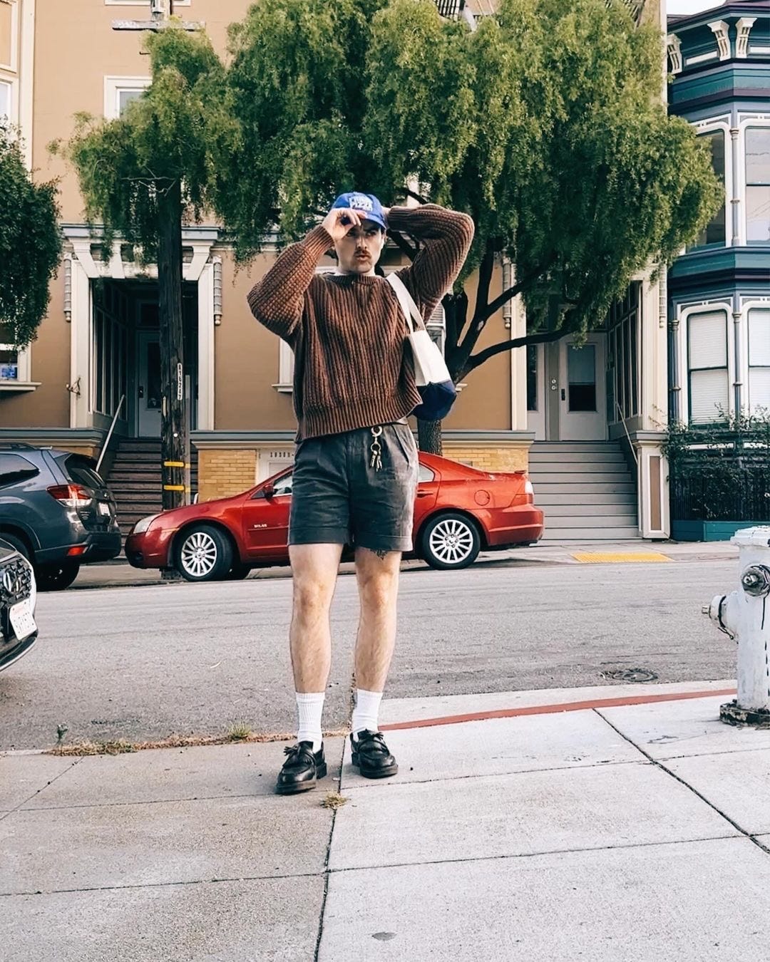 man standing on the sidewalk wearing a brown sweater and navy shorts with white socks and black loafers