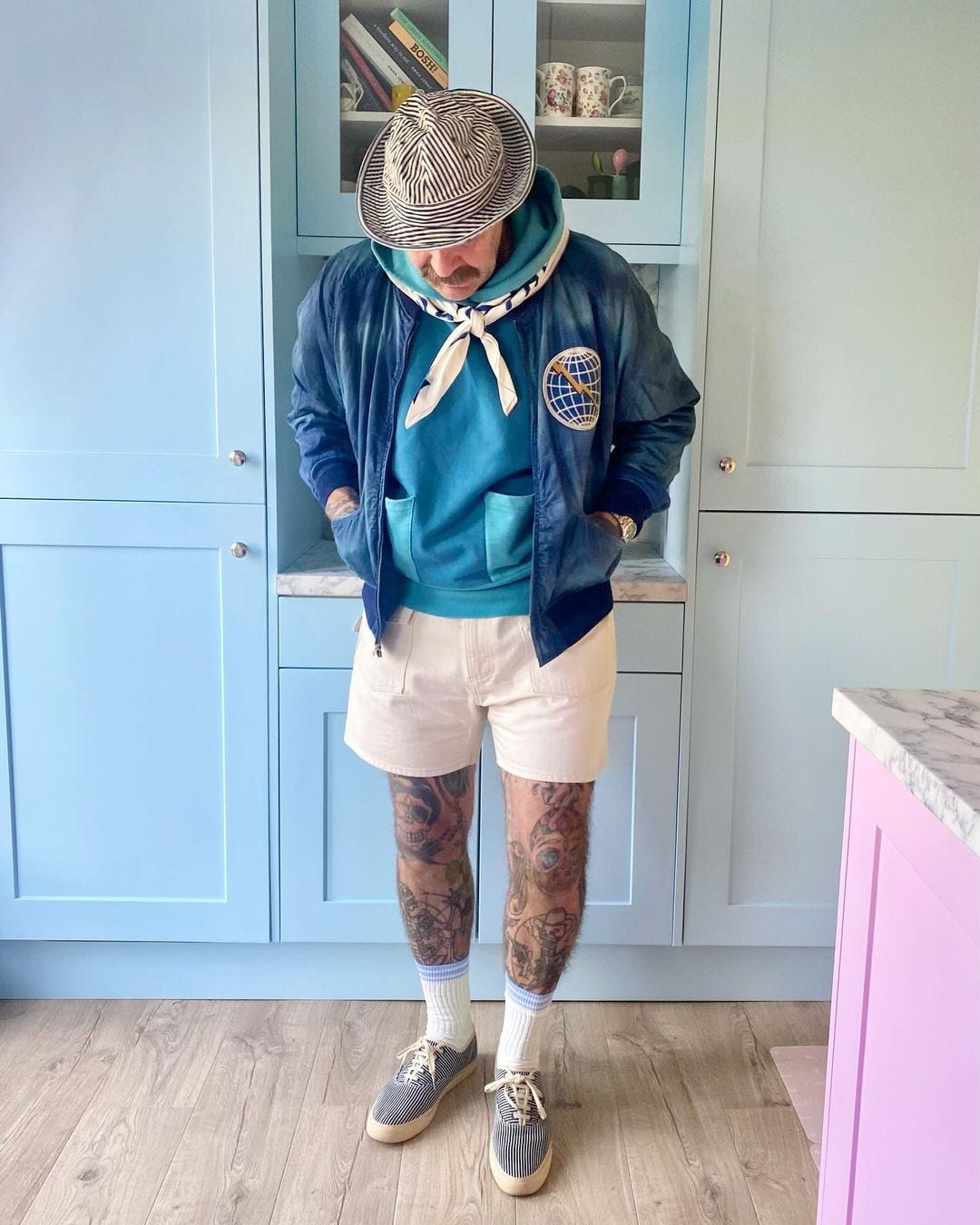 man standing in front of a cabinet, wearing a blue varsity jacket, light shorts, blue sneakers, and tall white socks