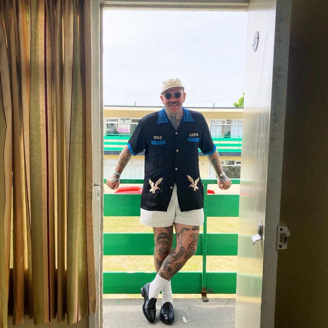 man standing on a patio wearing a short sleeve blue shirt, light shorts, white socks and loafers