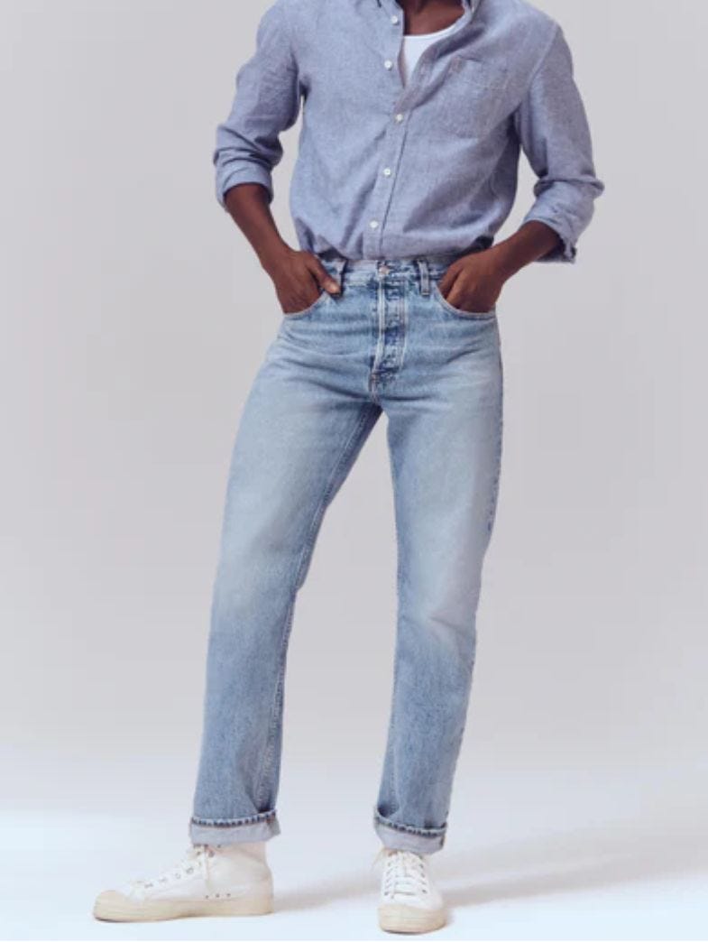man from the neck down wearing a light blue chambray shirt, light rinse jeans, and white high top sneakers