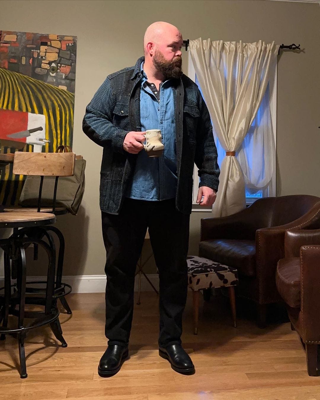 man standing in a kitchen holding a coffee mug, wearing a navy and black overshirt over a denim shirt and henley shirt with dark pants