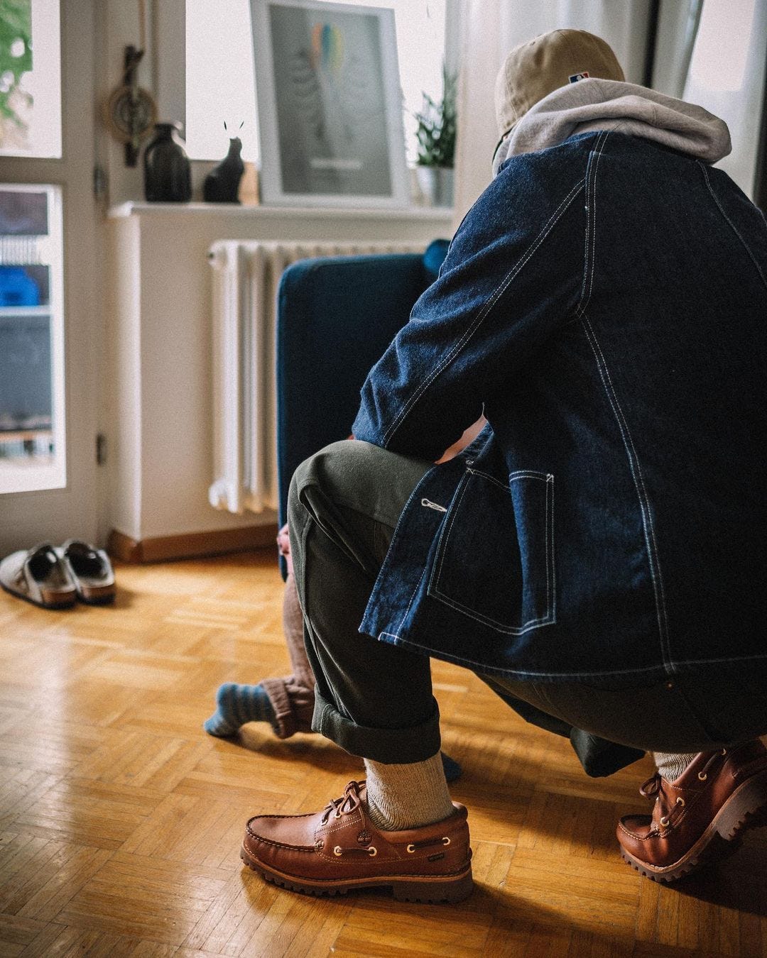 a man from behind, crouching on the floor wearing a denim jacket, olive green pants, and brown boat shoes
