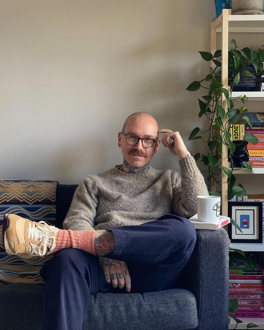 stylish man wearing glasses, a heather crewneck sweater, blue pants, and red socks with new balance sneakers