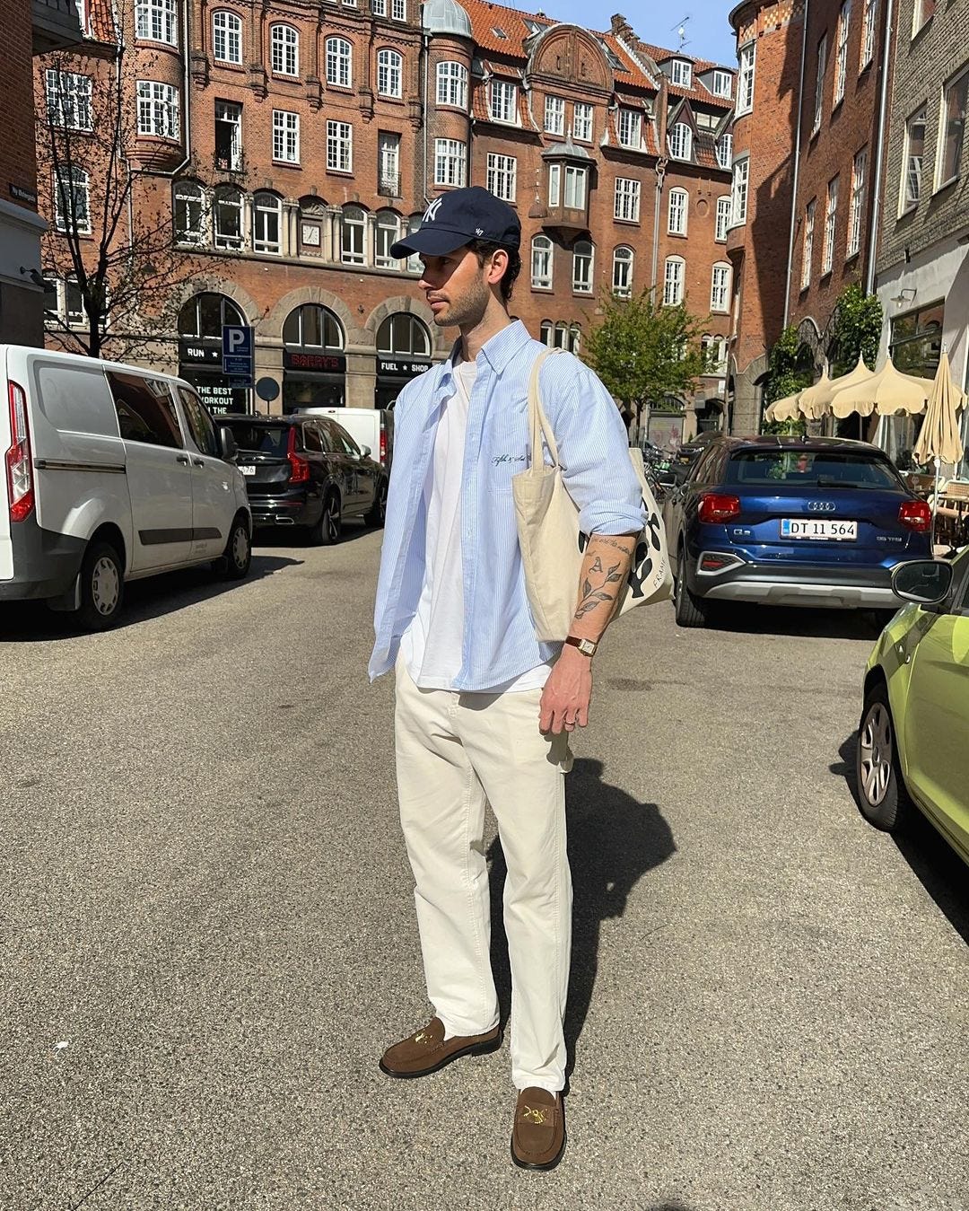 man standing in the street, wearing a light blue button-down shirt over a white t-shirt, light-colored pants, and brown loafers