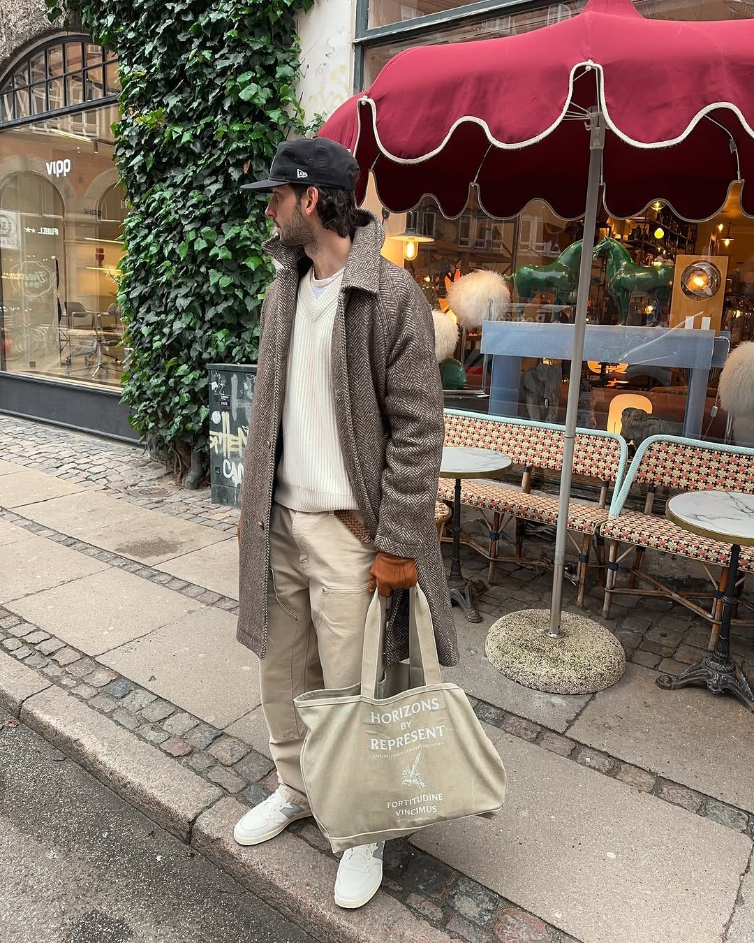 stylish man wearing herringbone topcoat, cream-colored crewneck sweater, tan chinos, and white New Balance sneakers