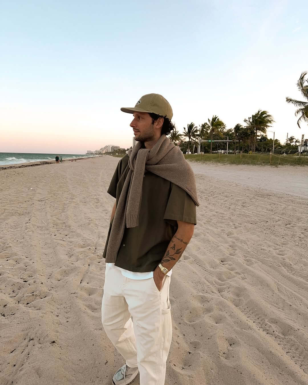 stylish man standing on a beach wearing a brown sweater tied around his neck over a brown short sleeve button-down shirt with white jeans and sneakers