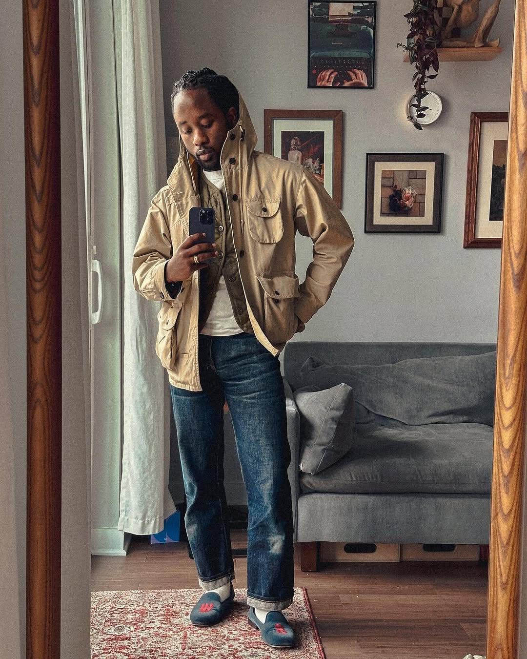 stylish man taking a selfie in front of a grey couch wearing a tan field jacket and dark blue jeans