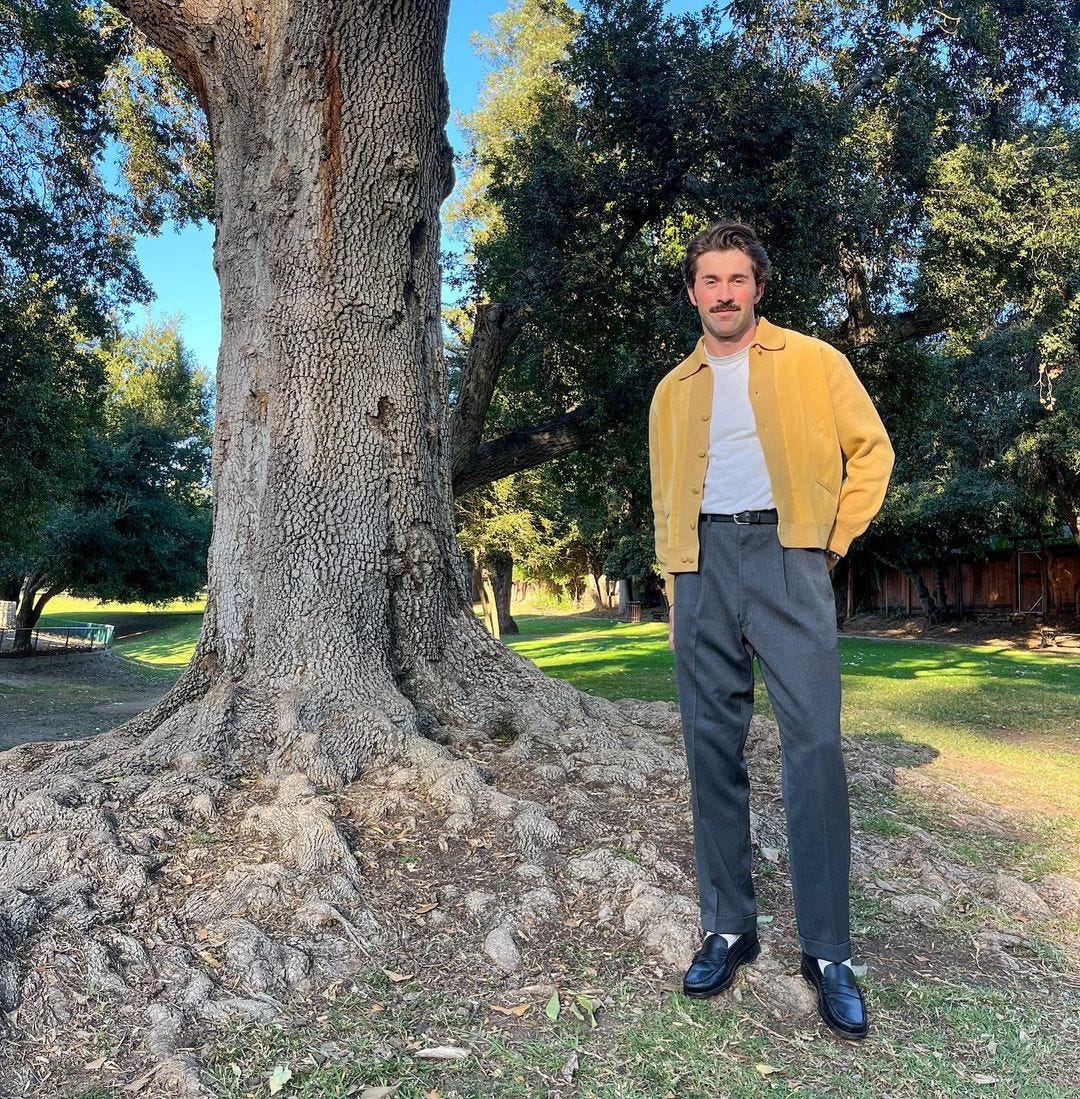 stylish man wearing a yellow collared cardigan over a white t-shirt with grey trousers and a black belt