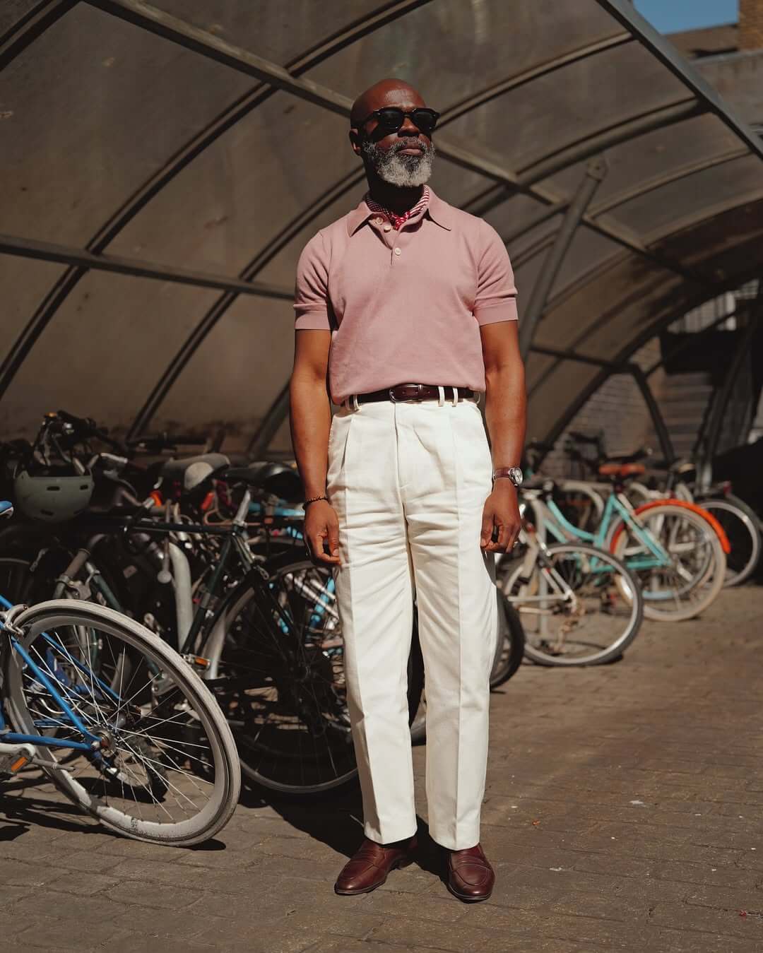 stylish man wearing a dusty rose knit polo with crisp white trousers, a brown belt, red bandana, and brown shoes