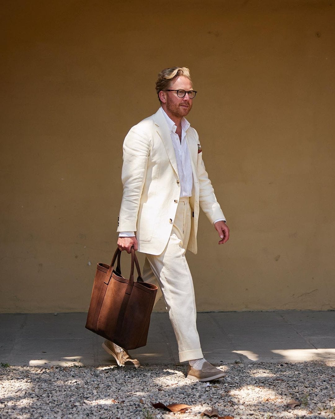 stylish man wearing an off-white summer suit with a white shirt and tan loafers, carrying a brown leather bag