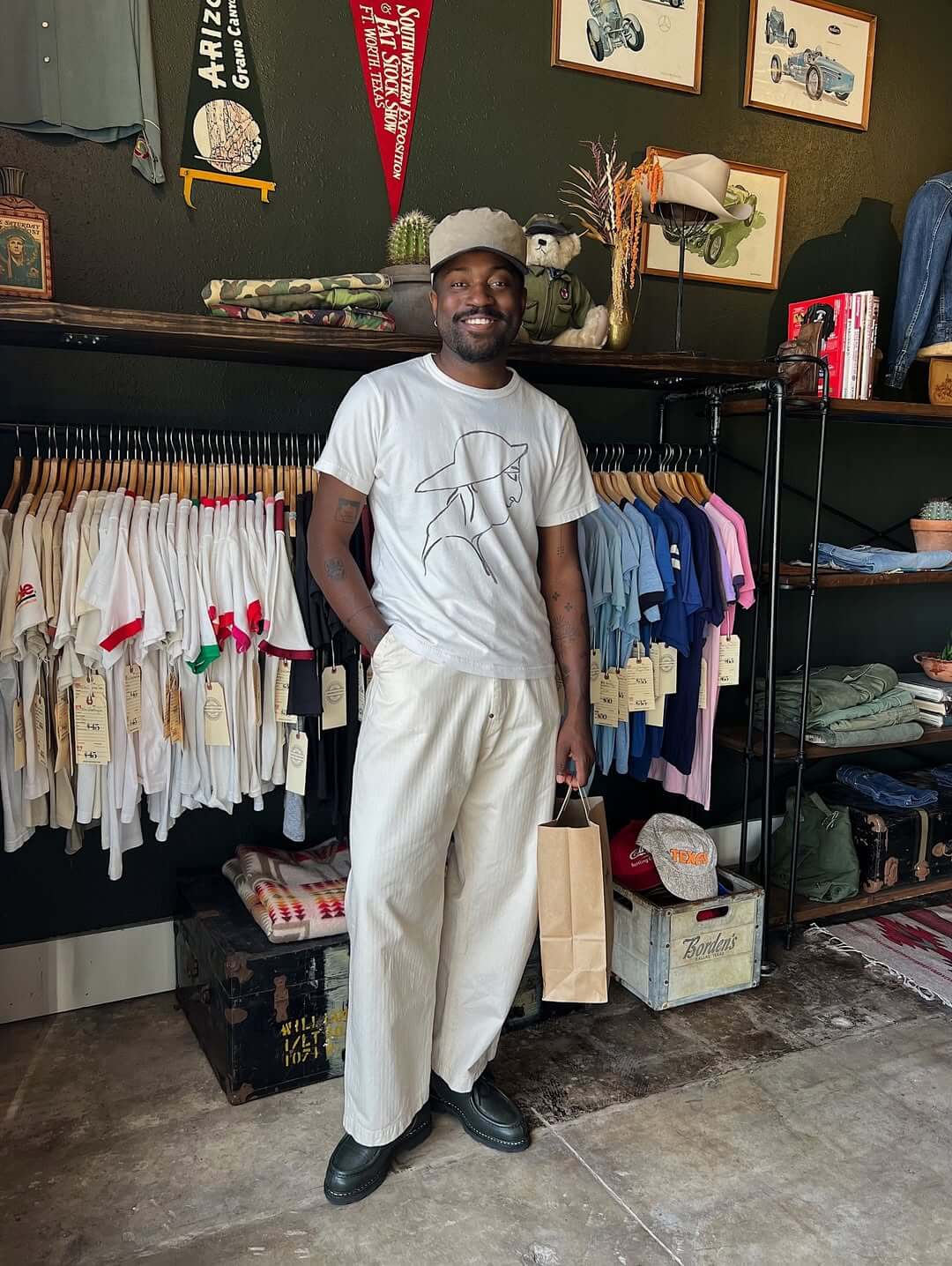 stylish man standing in a clothing store wearing a white graphic tee, white pants, and black shoes