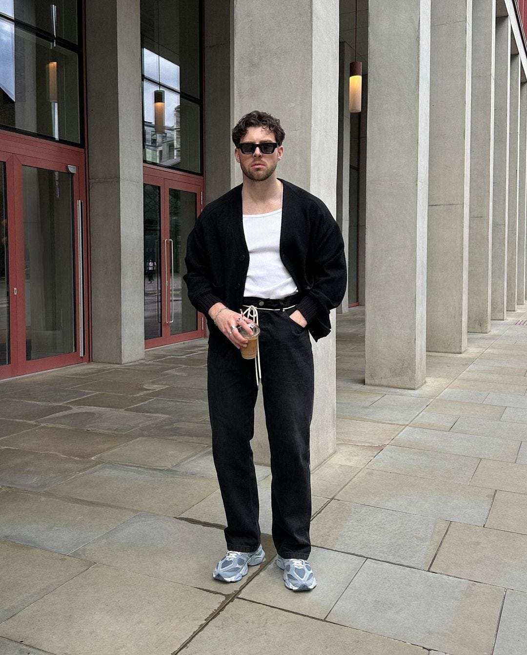 man standing outside in sunglasses, wearing a black cardigan, white tank top and black drawstring pants. he's holding an iced coffee