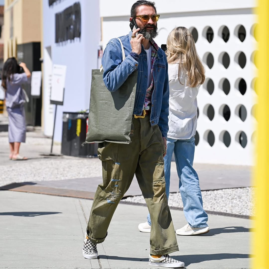 stylish man walking down the street wearing olive-colored pants, a denim jacket and checkerboard vans sneakers