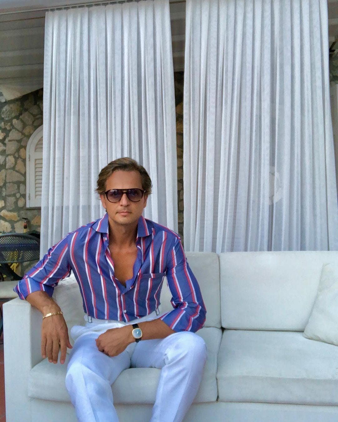 stylish man seated on a white couch wearing a blue and red striped shirt and white pants with sunglasses