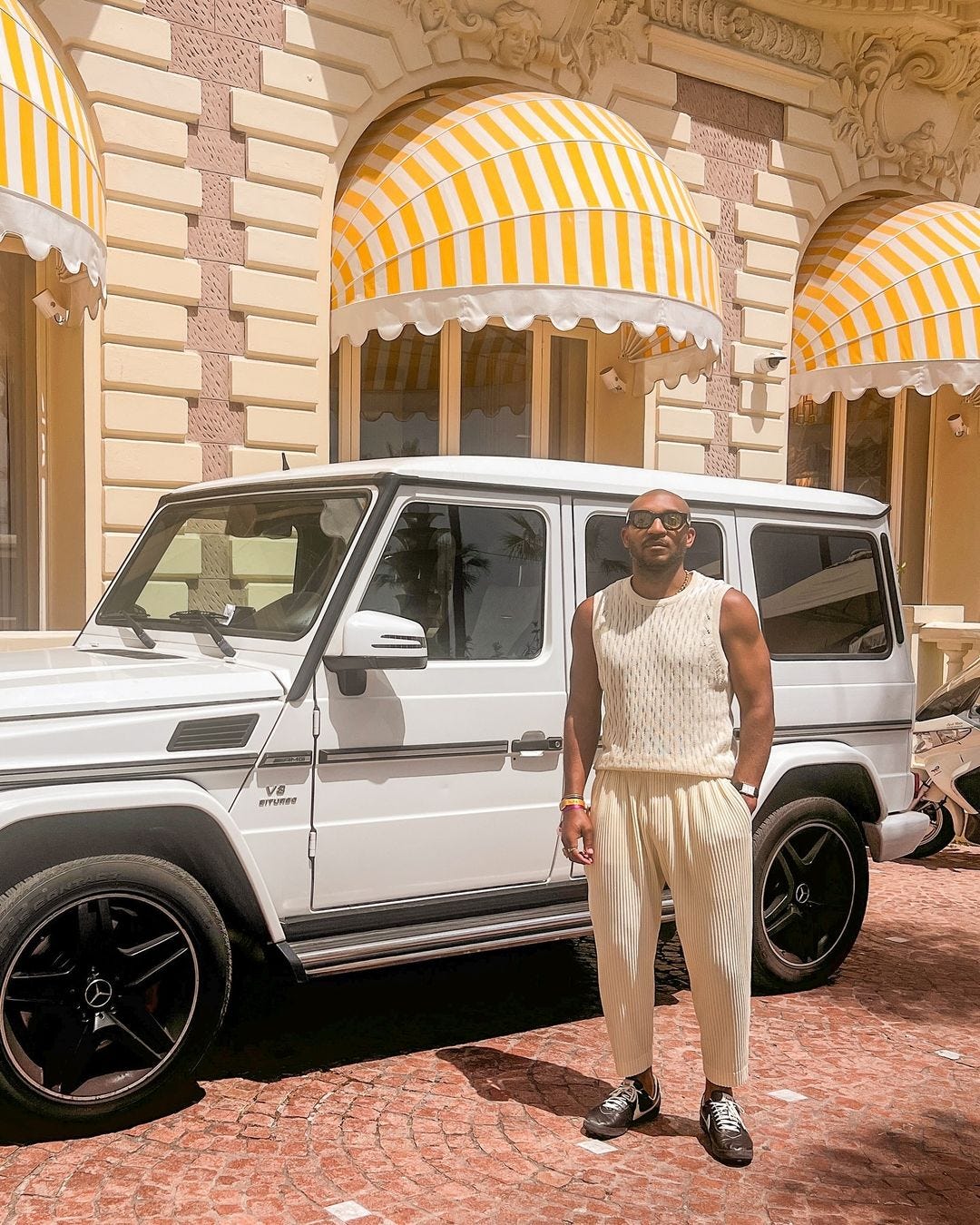 a man standing outside in the sun in front of a white car, wearing a crochet sleeveless top, casual pants and sneakers, light colored outfits for men