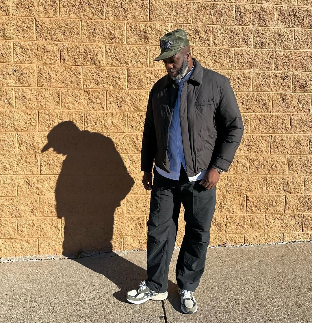 stylish man wearing a button down shirt, black coach's jacket, dark pants, and New Balance sneakers