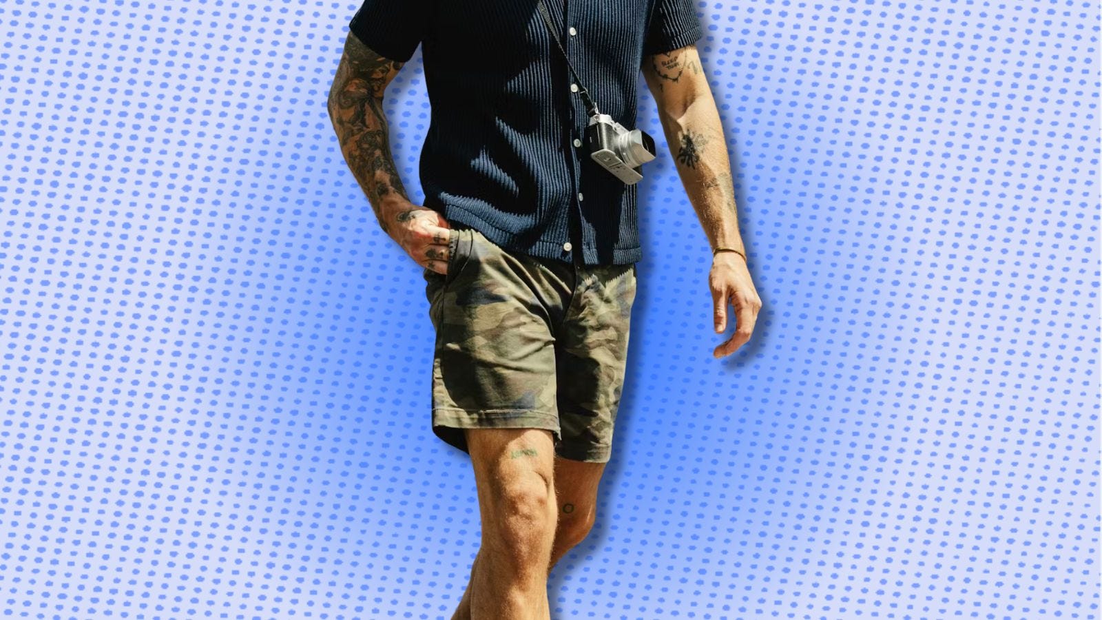 man wearing a navy sweater polo, camo shorts, and a camera across his chest