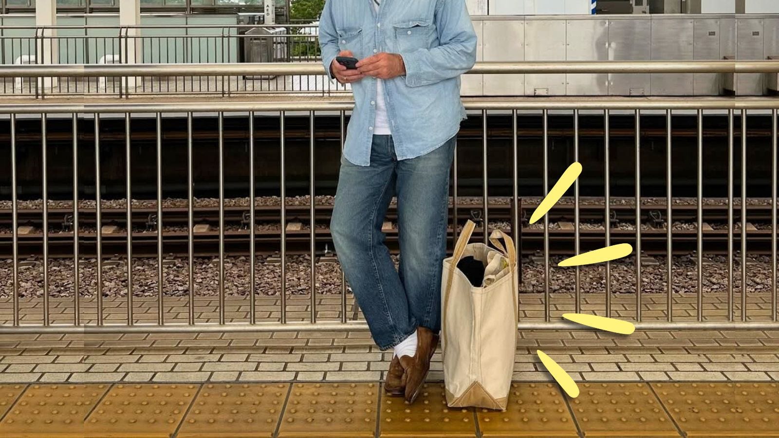 stylish man from the neck down wearing a chambray shirt, jeans, loafers, with a canvas tote bag at his feet