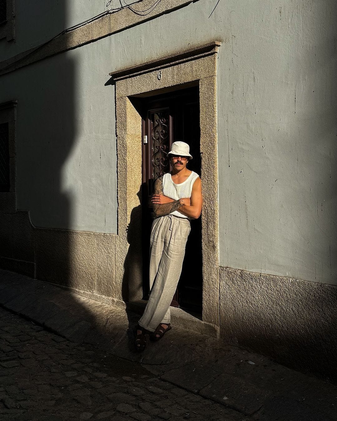 man standing in a doorway outside, wearing a white bucket cap, knit sleeveless shirt, and tan linen pants