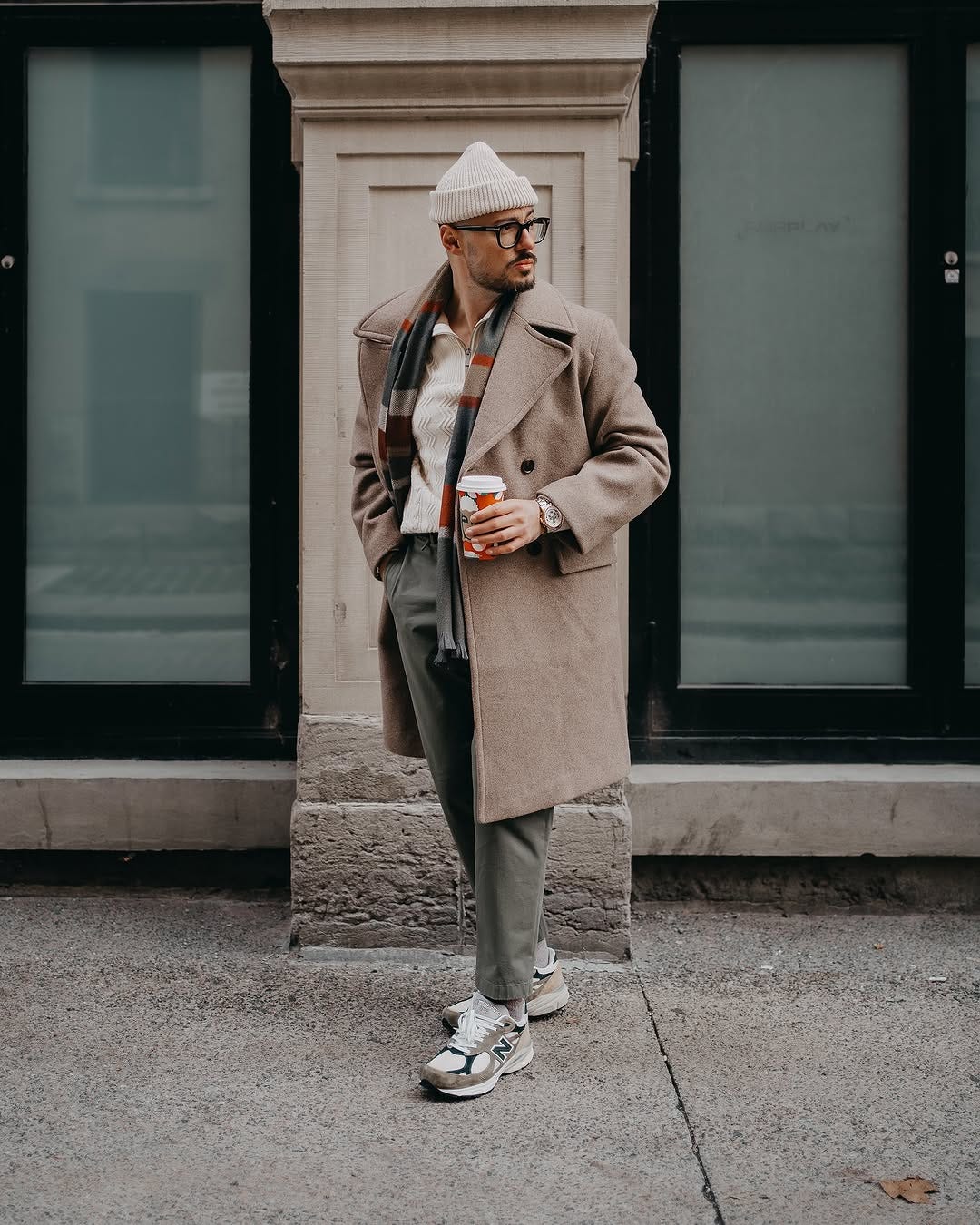stylish man wearing a quarter zip, plaid scarf, olive green chinos, a tan topcoat, and new balance sneakers