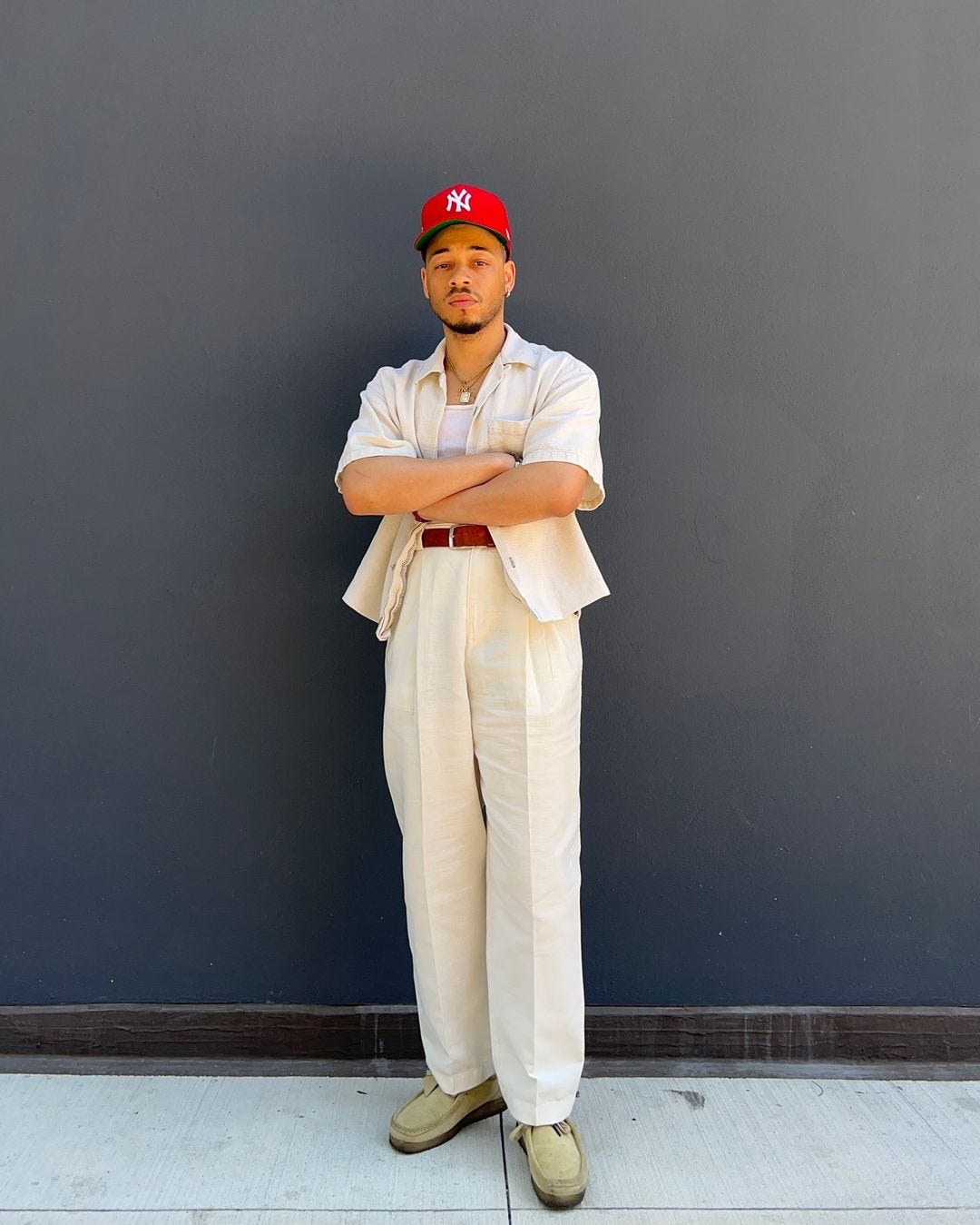 man wearing a light colored short-sleeve shirt over a white undershirt, with light colored pants and suede shoes. he has his arms crossed and has a red baseball cap on his head