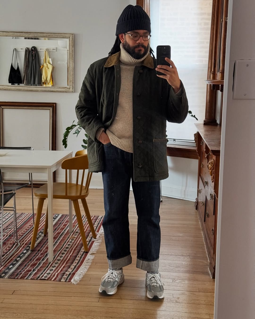 stylish man wearing an oatmeal-colored turtleneck, black beanie, green barn jacket, dark jeans, and grey New Balance 99ov6 sneakers
