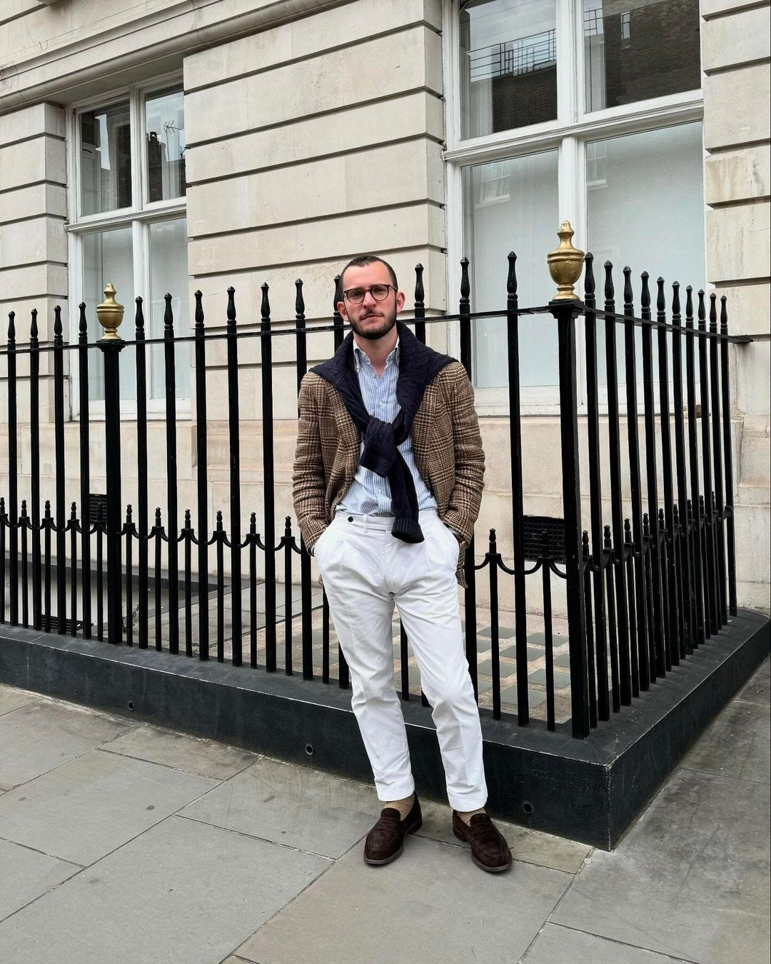 man standing on a street corner wearing a sport coat and chinos with a sweater over his shoulders, and loafers on his feet