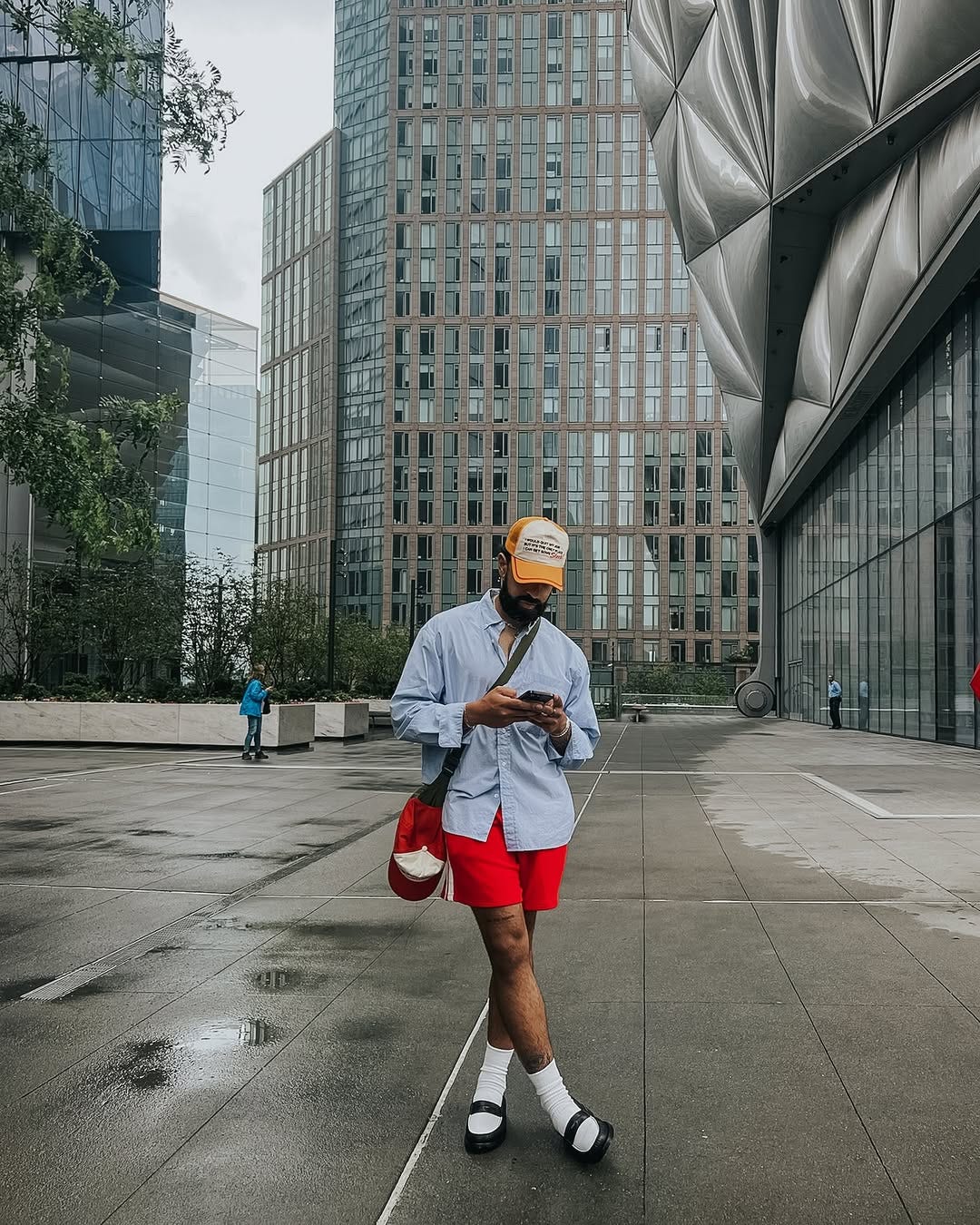 stylish man standing outside a large building wearing a blue oxford shirt, crossbody bag, red shorts, black and white loafers, and tall white socks