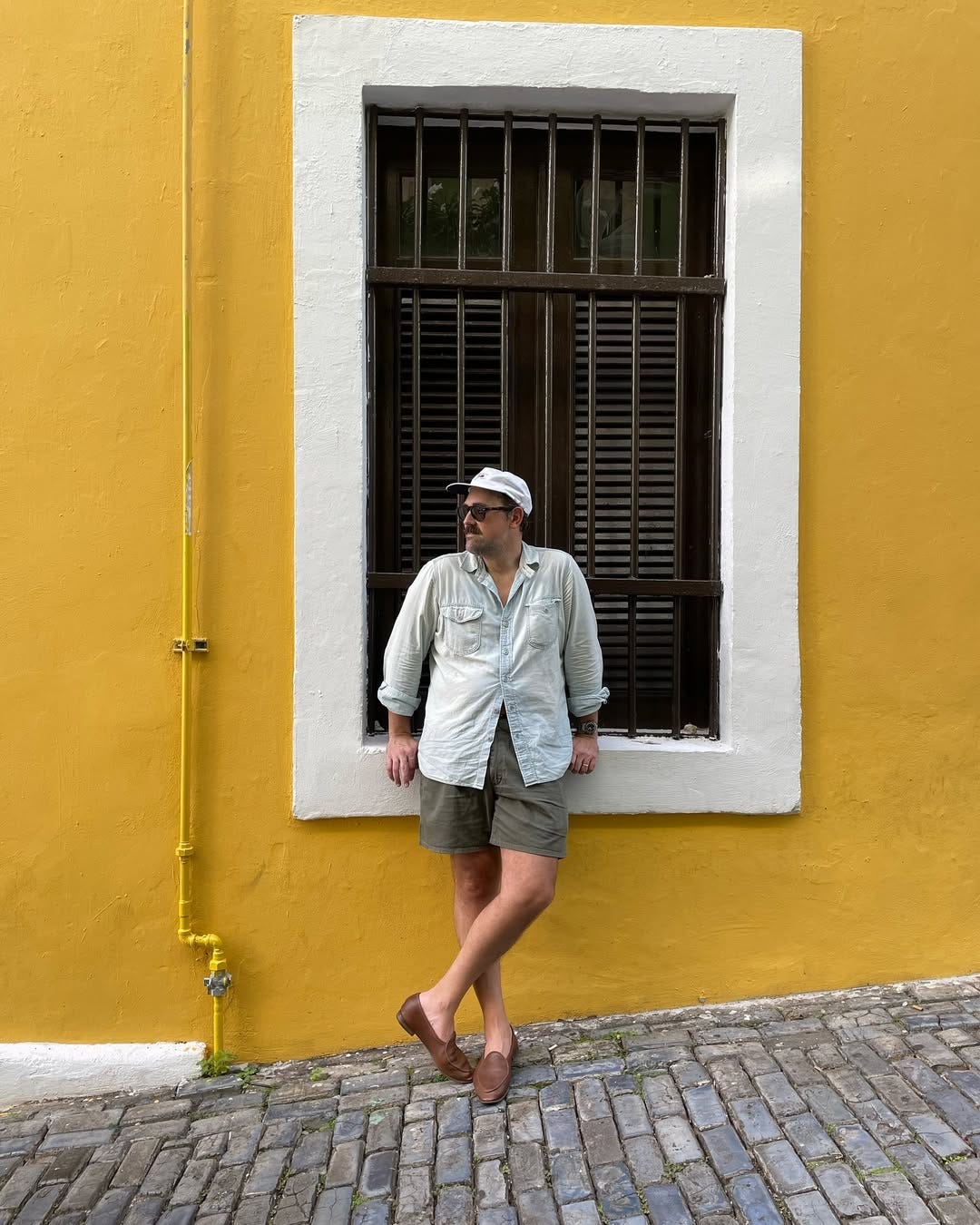 stylish man standing outside in front of a brightly painted wall wearing a chambray shirt, grey shorts, and brown loafers