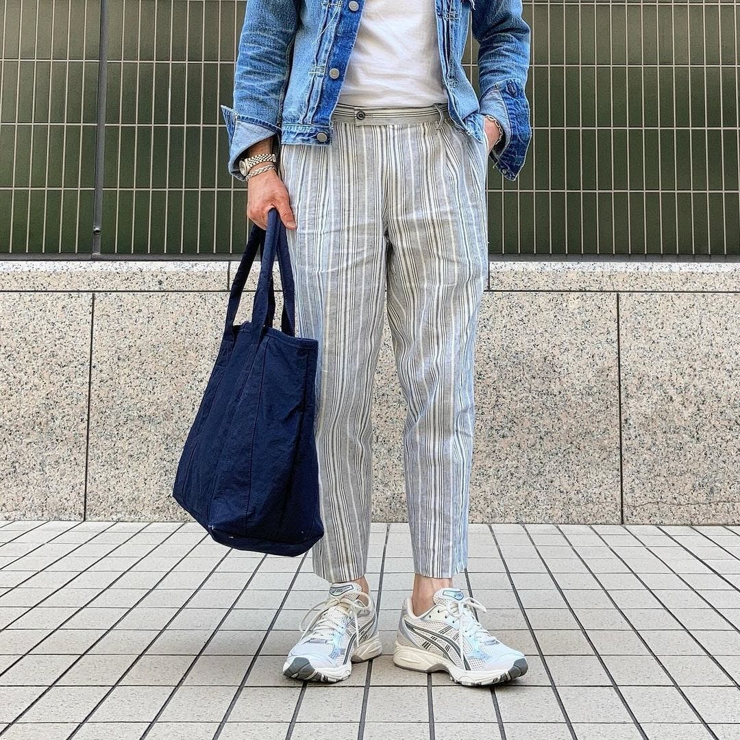 man wearing glasses, a denim jacket, white t-shirt, and striped pants, holding a navy bag