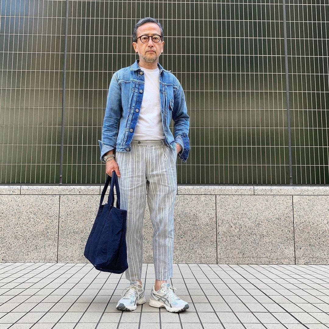 man in a denim jacket, white t-shirt, and striped pants, holding a navy bag