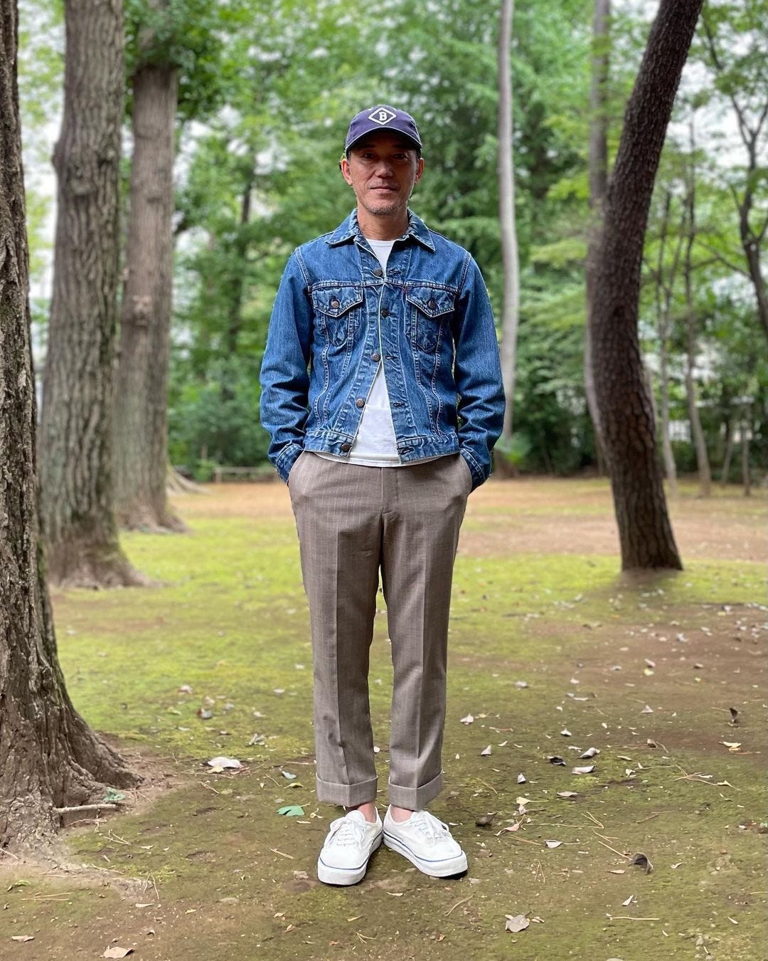 man standing among trees wearing a denim jacket with brown pants and white sneakers