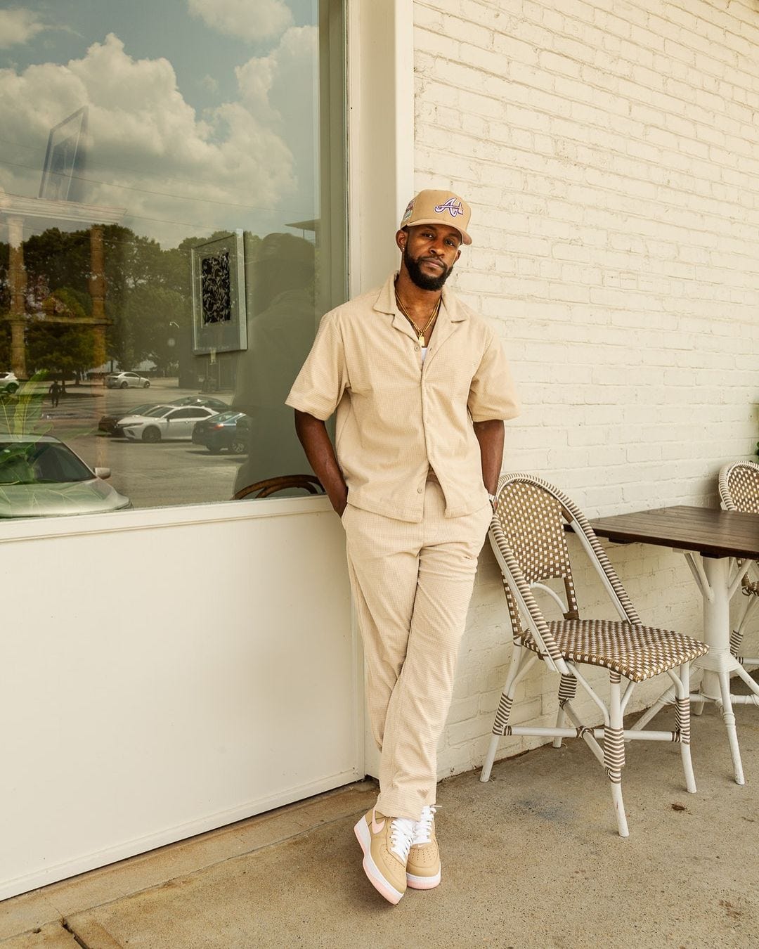 man standing outside of a cafe wearing a tan short sleeve shirt with matching tan pants. he has his hands in his pockets and is looking directly at the camera. his legs are crossed