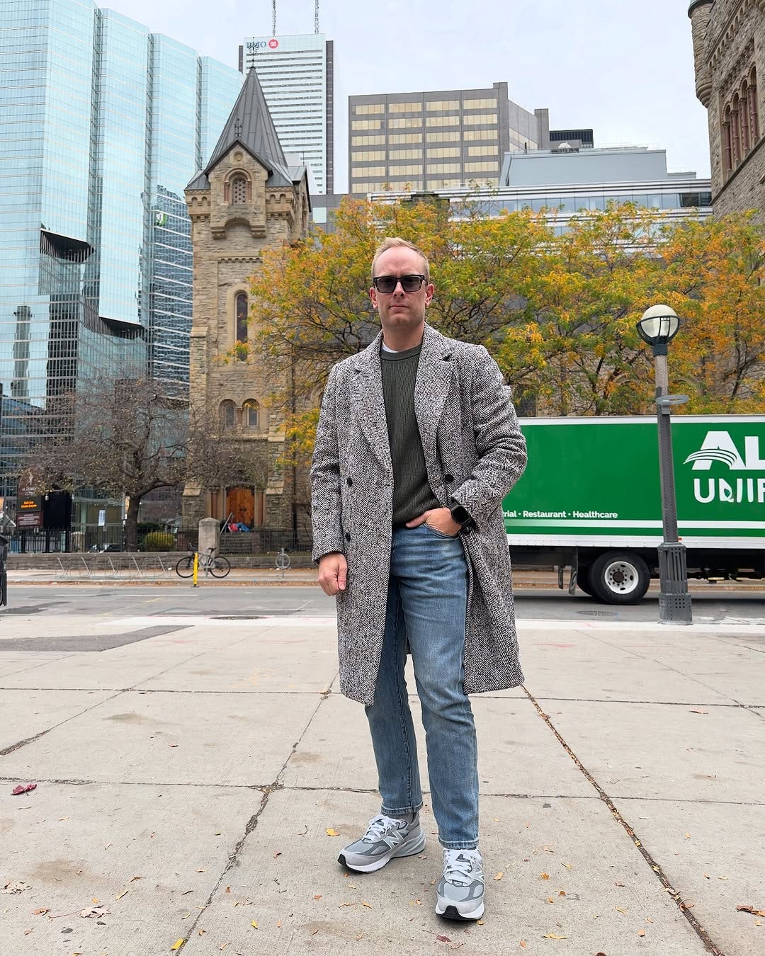 stylish man wearing a patterned topcoat over a grey crewneck sweater, blue jeans, and gray New Balance 99ov6 sneakers