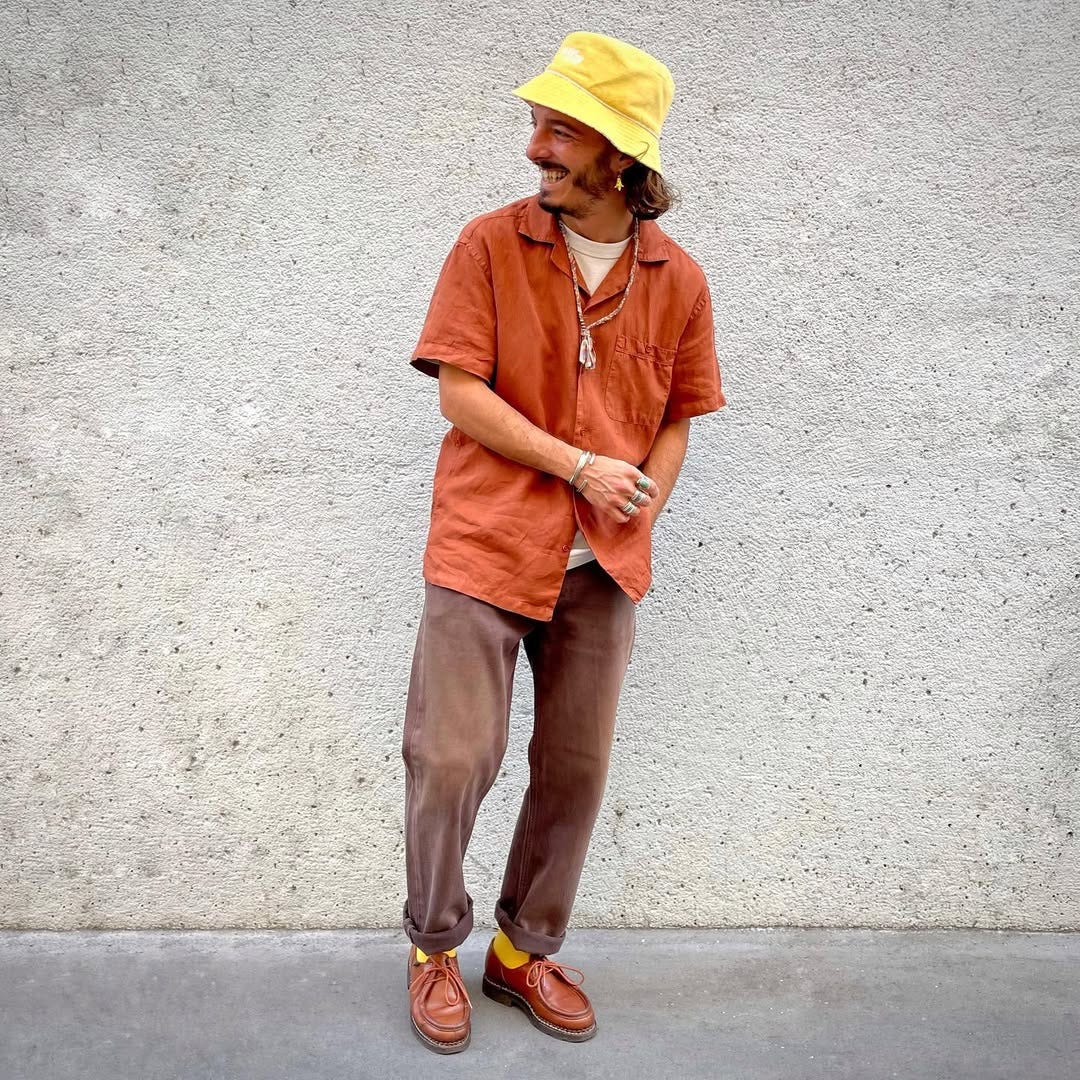 stylish man wearing an orange camp shirt, yellow bucket hat, and brown pants