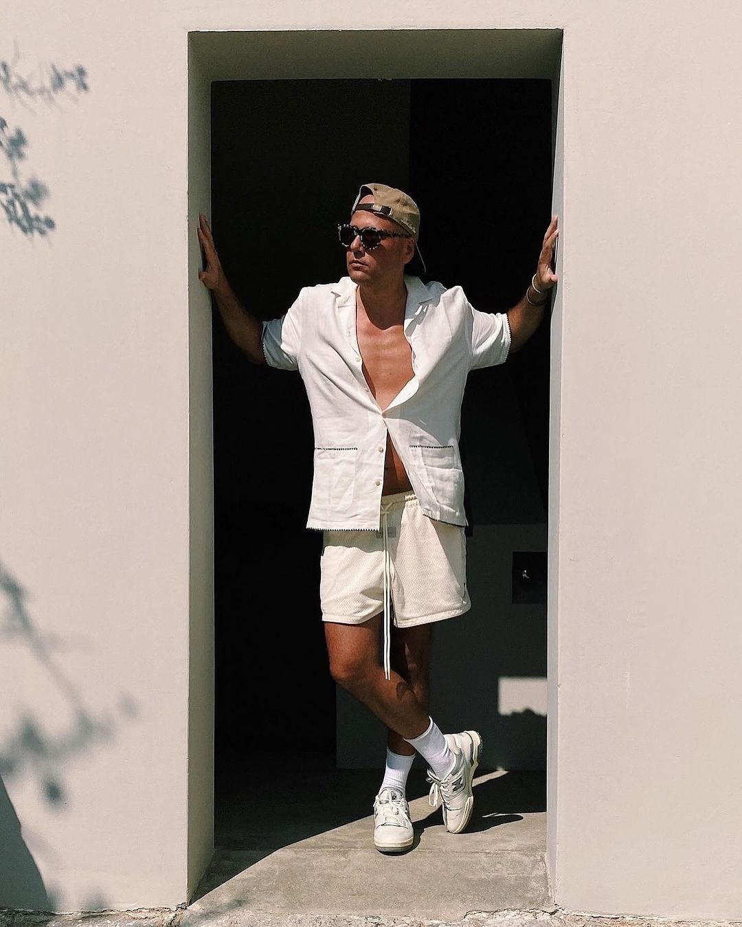man in a doorway wearing a short-sleeve white shirt and white drawstring shorts with white sneakers and tall socks