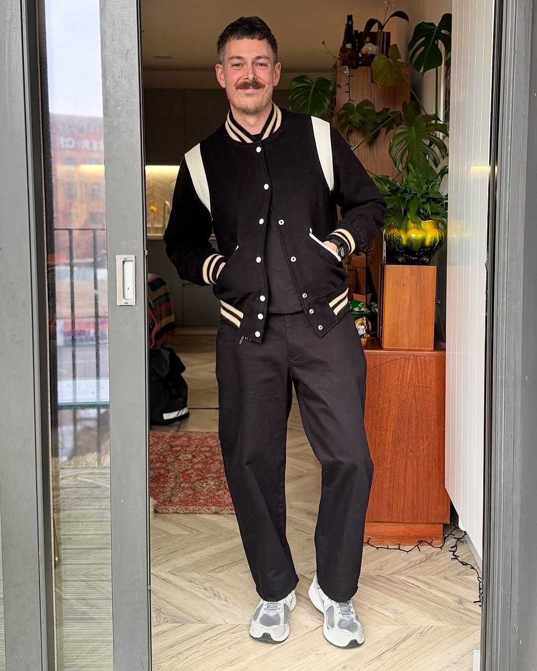 stylish man standing in a doorway with his hands in the pockets of a black letterman jacket over a black crewneck sweatshirt, with black trousers and New Balance 99ov6 sneakers