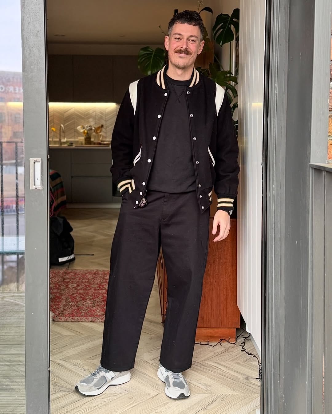 stylish man wearing a black letterman jacket over a black crewneck sweatshirt, with black trousers and New Balance 99ov6 sneakers