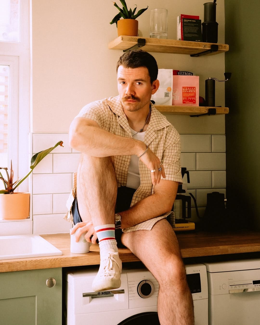 stylish man sitting on a kitchen counter wearing a camp shirt, shorts, and athletic socks